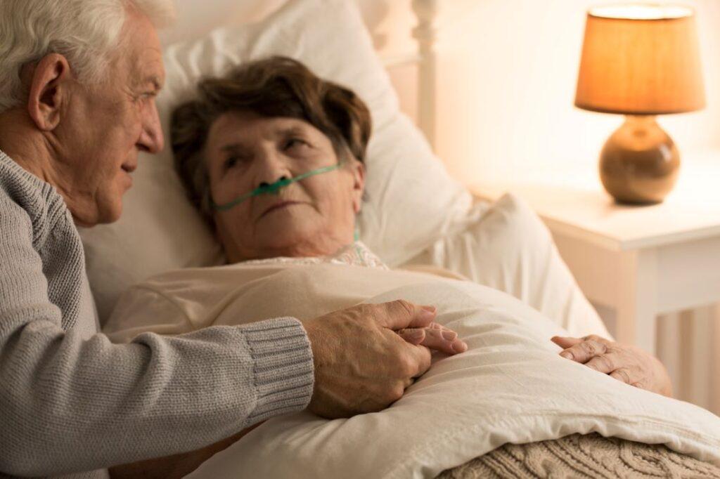 Elderly man, support sick wife while she lays in bed, holding his hand.